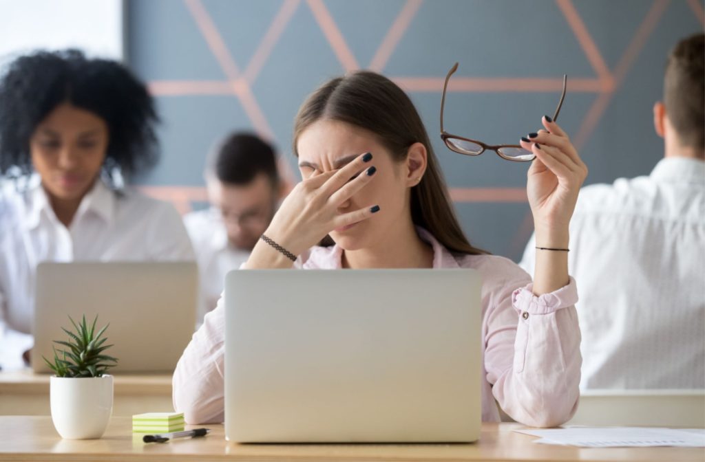 A woman sitting at a desk while she takes off her glasses and rubs her eye due to eye strain from her computer