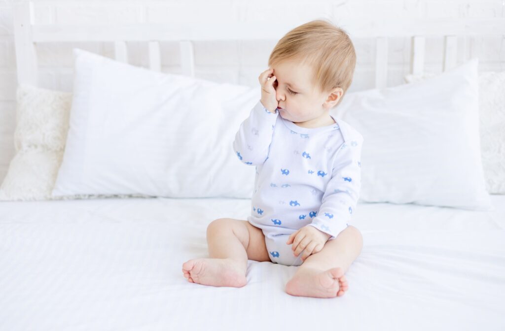 A baby sitting upright on a white bed, rubbing their eyes in frustration.