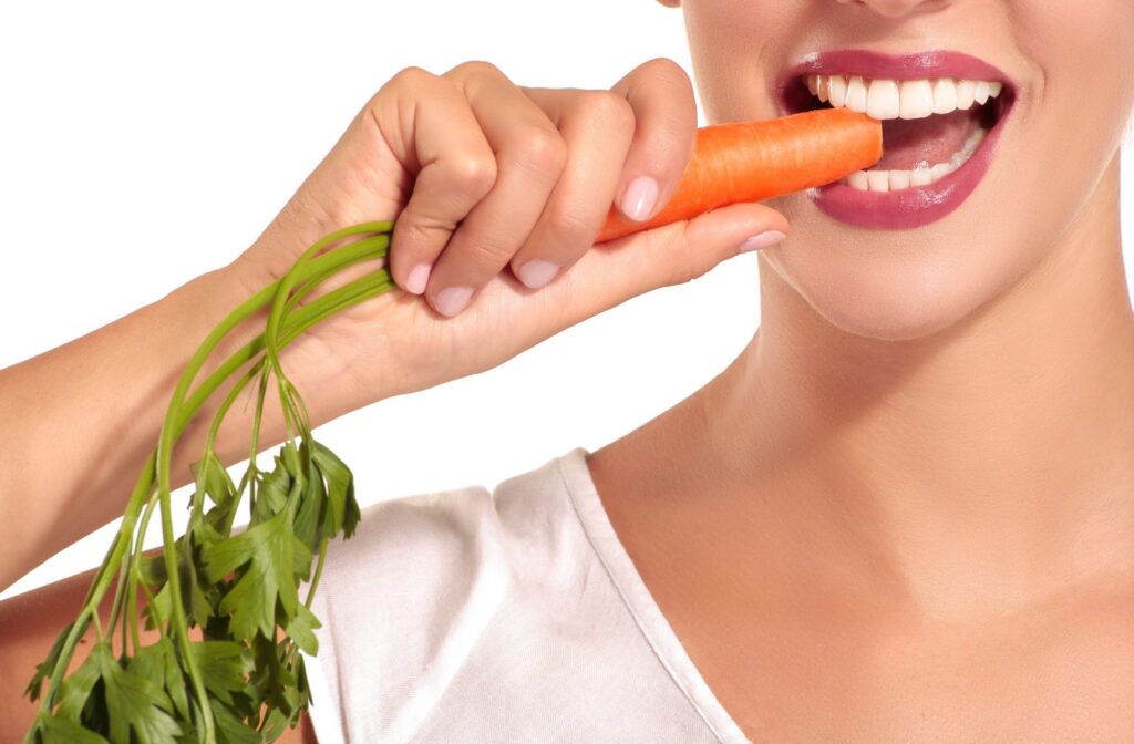 A close-up image of a person taking a bite from the end of a carrot in front of a white background.