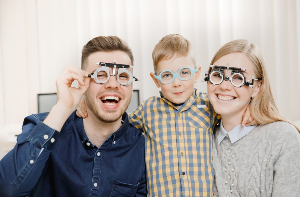 Two laughing parents on either side of their smirking young child as all wear instruments for testing vision at an eye exam