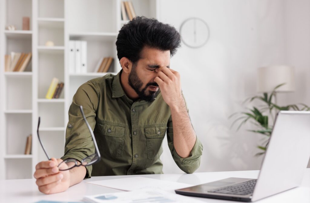 A person sitting at their desk with a laptop, hands placed over eyes indicating eye pain