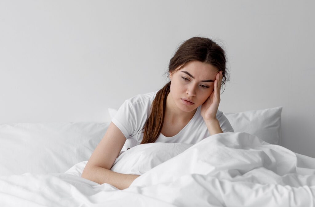 A young adult sitting in bed with a hand on their head expressing discomfort from a headache