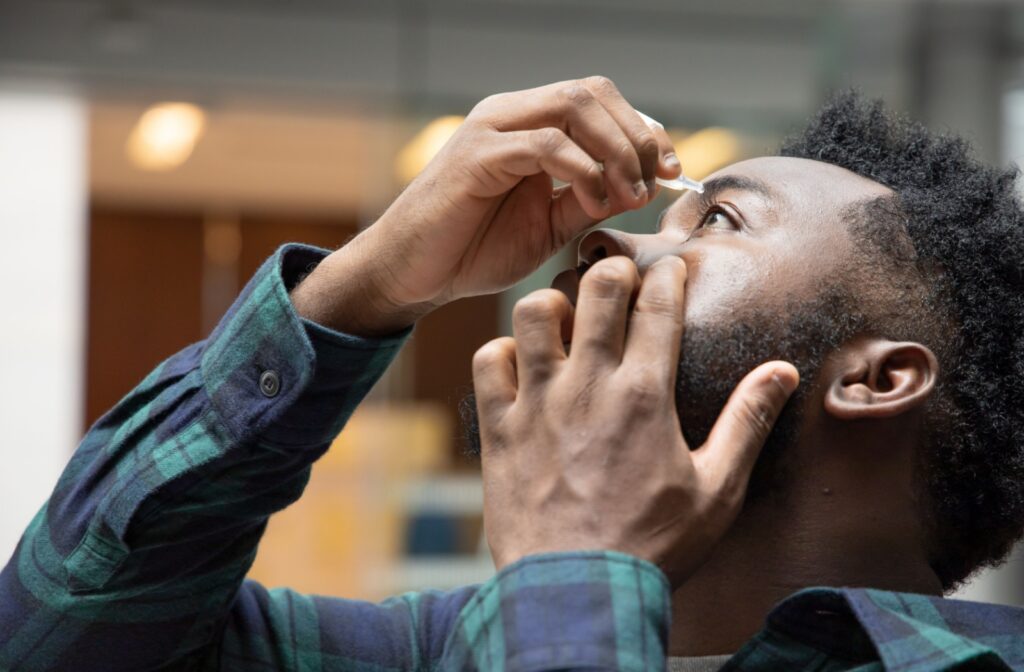Man in dark green plaid shirt instilling eye drops into left eye.