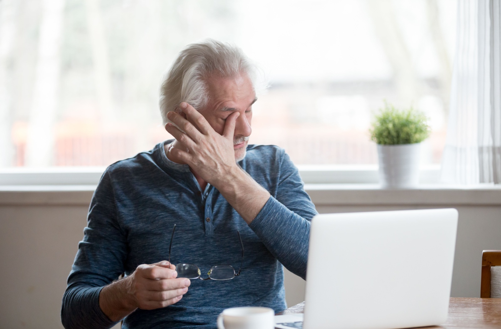 An person sits at a table in front of a laptop, holding his glasses in one hand and rubbing his eye with the other.
