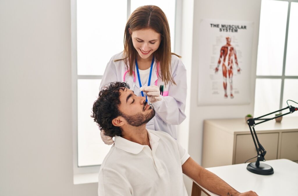 A smiling doctor applies eye drops to a seated patient during an eye exam in a medical office.
