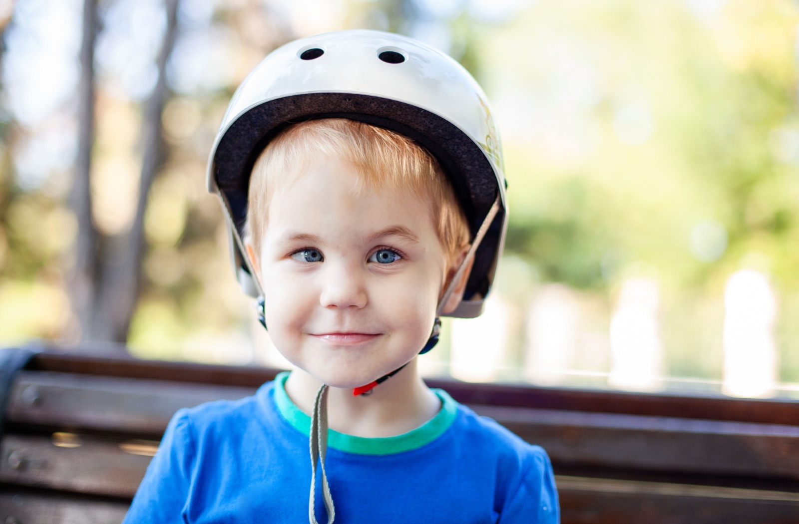 Young boy in white helmet with crossed eyes sitting on a bench outdoors.