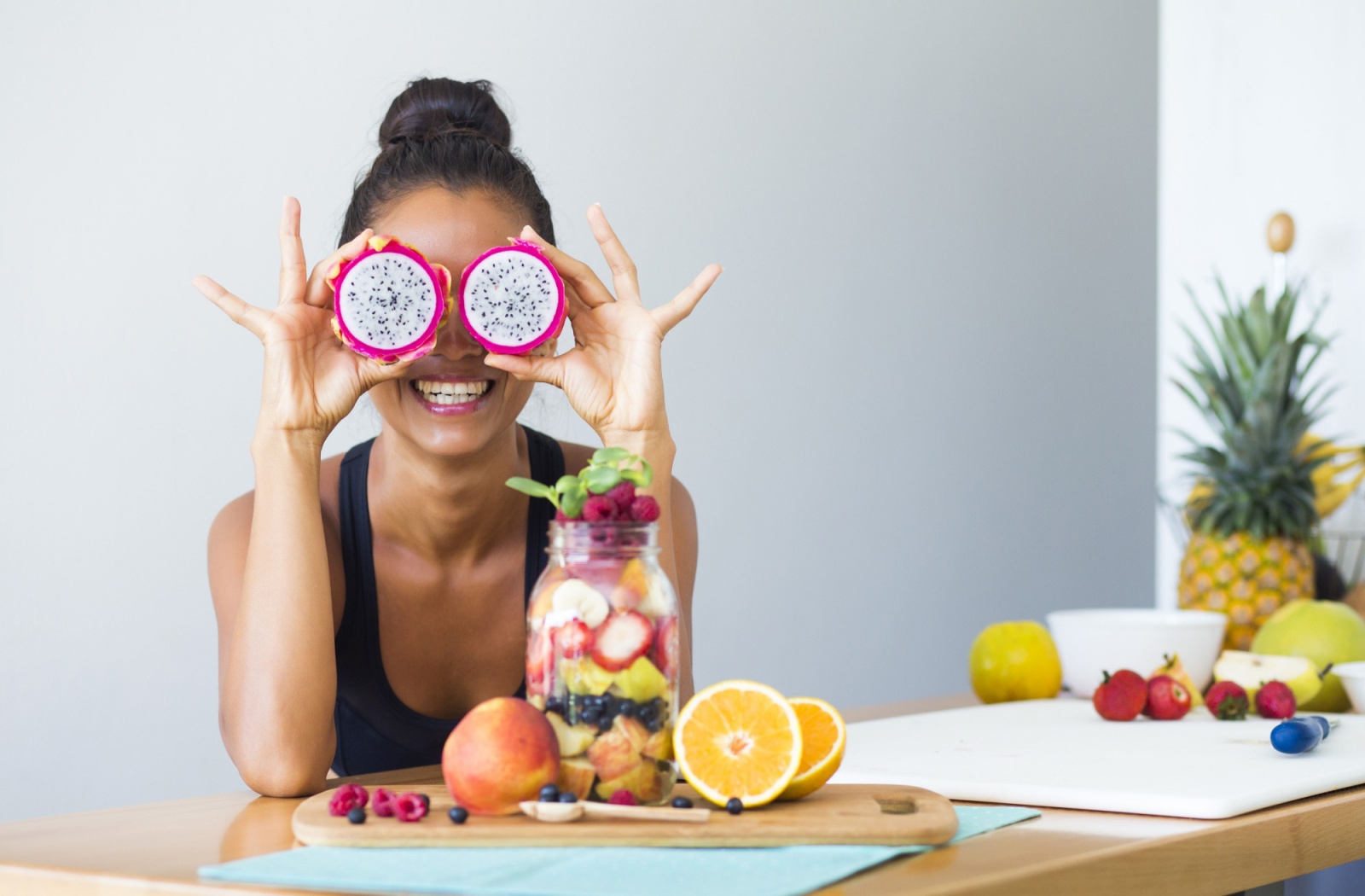 A person leaning on a counter top covered in fruit, holding up 2 pieces of dragon fruit in front of their eyes.